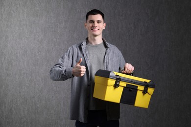 Young man with tool box showing thumbs up on grey background