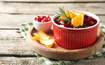 Photo of Cranberry sauce in bowl, fresh berries, rosemary and orange peels on wooden table, closeup. Space for text