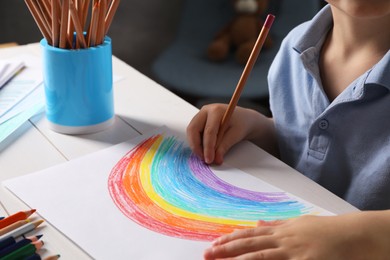 Photo of Little boy drawing rainbow with pencil at white wooden table indoors, closeup. Child`s art