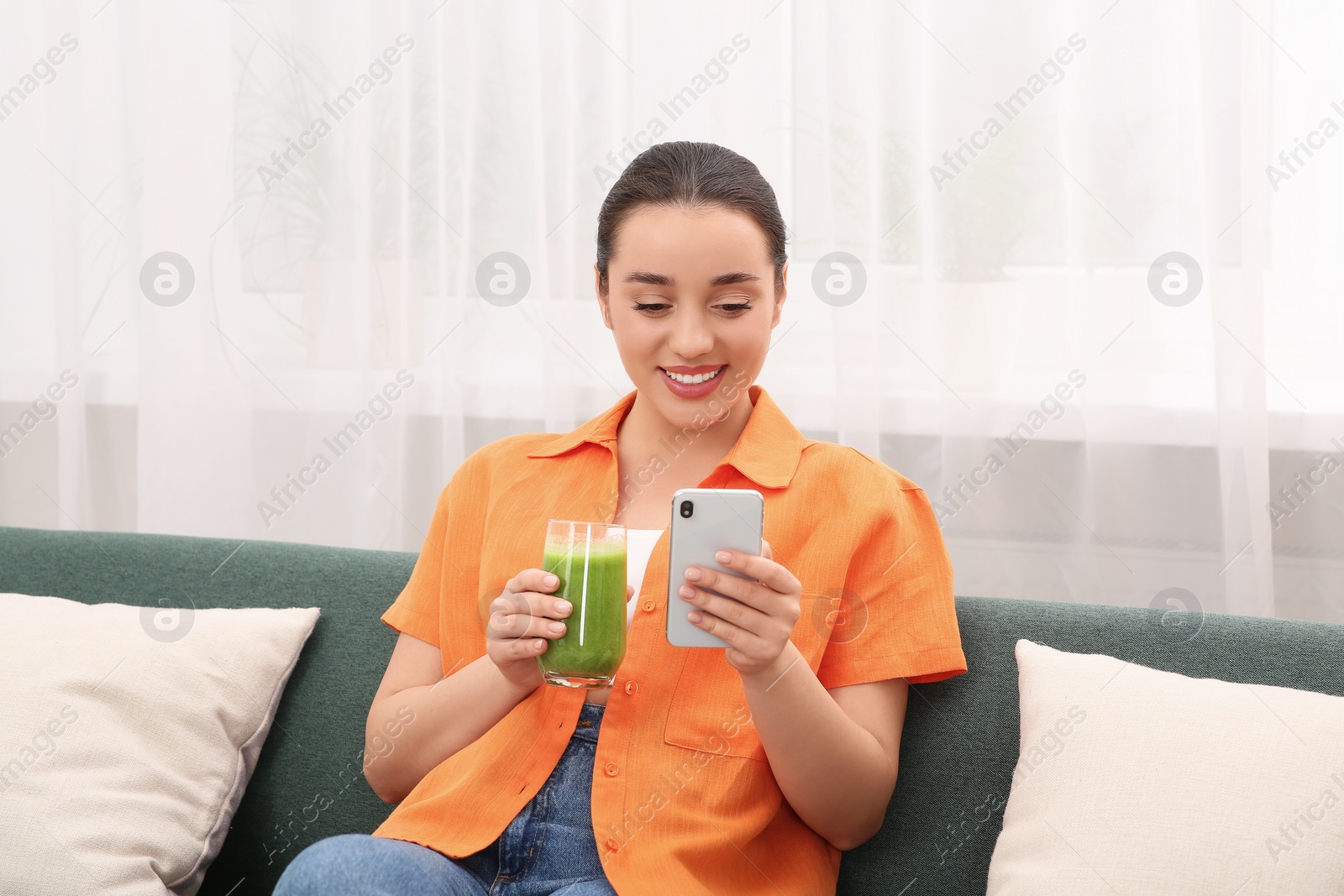 Photo of Beautiful young woman with glass of delicious smoothie using smartphone indoors