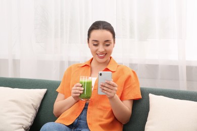 Photo of Beautiful young woman with glass of delicious smoothie using smartphone indoors