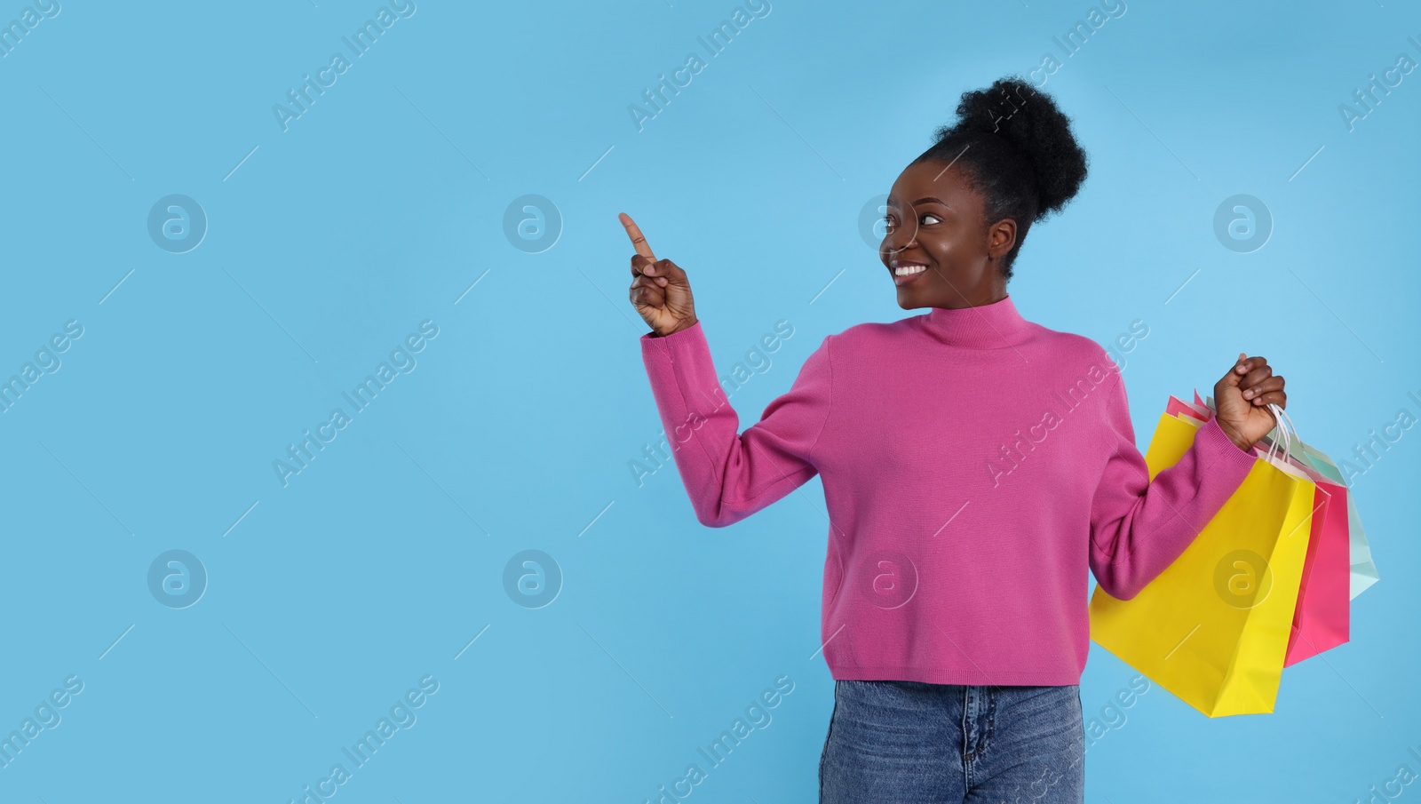 Photo of Happy young woman with shopping bags pointing at something on light blue background. Space for text