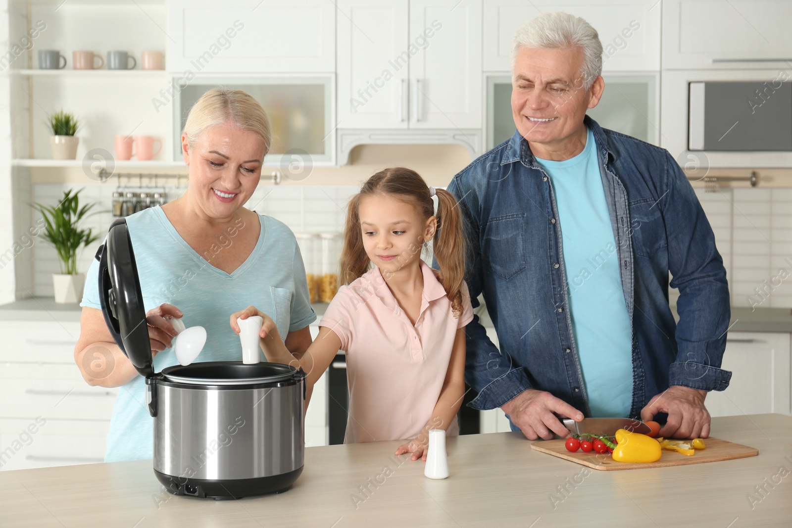Photo of Mature couple and their granddaughter preparing food with modern multi cooker in kitchen