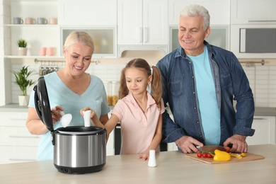 Mature couple and their granddaughter preparing food with modern multi cooker in kitchen