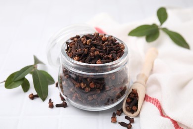 Photo of Aromatic cloves in glass jar, scoop and green leaves on white table, closeup