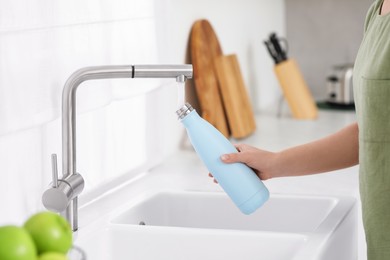 Photo of Woman pouring fresh water from tap into thermo bottle in kitchen, closeup
