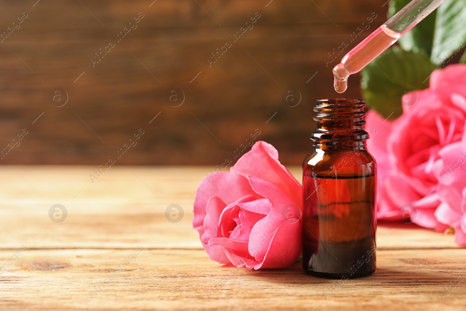 Photo of Bottle of rose essential oil, pipette and flowers on wooden table, space for text