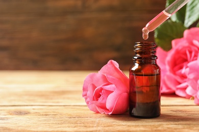 Photo of Bottle of rose essential oil, pipette and flowers on wooden table, space for text