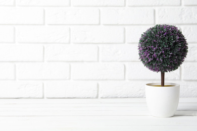 Photo of Beautiful artificial plant in flower pot on white wooden table near brick wall. Space for text