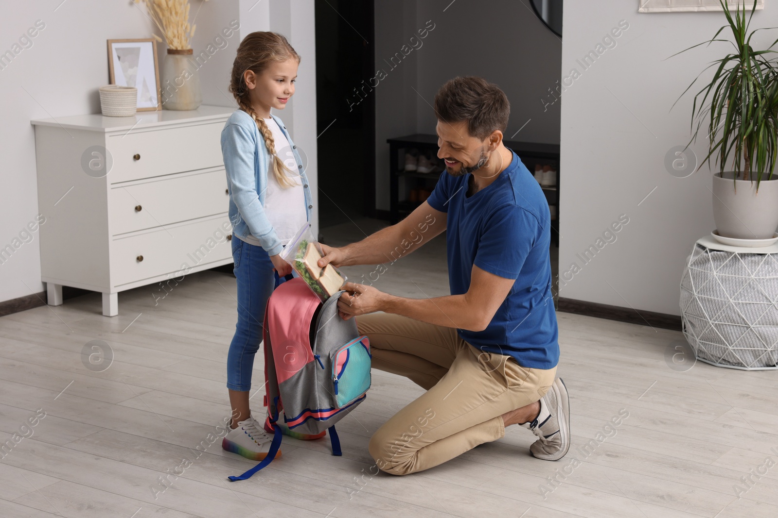 Photo of Happy father putting lunch box into daughter`s backpack at home. Preparing to school