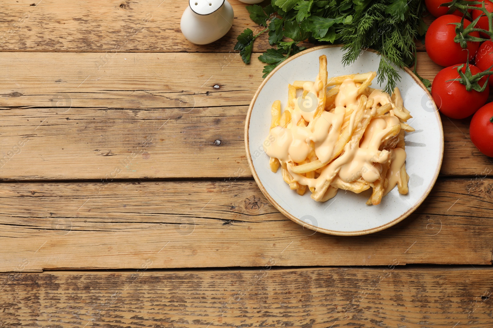 Photo of Delicious French fries with cheese sauce, tomatoes, dill and parsley on wooden table, top view. Space for text