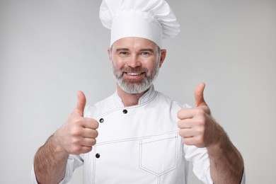 Happy chef in uniform showing thumbs up on grey background