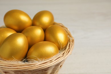 Photo of Shiny golden eggs in wicker basket on light background, closeup. Space for text