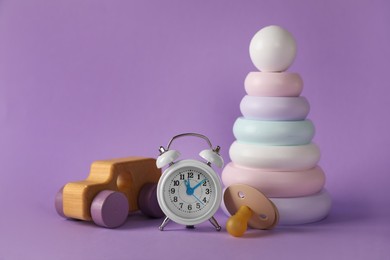 Alarm clock, toys and baby dummy on violet background. Time to give birth