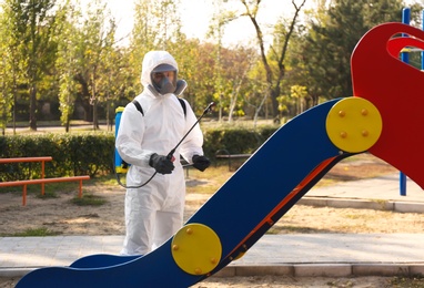 Photo of Man in hazmat suit with disinfectant sprayer near slide at children's playground. Surface treatment during coronavirus pandemic