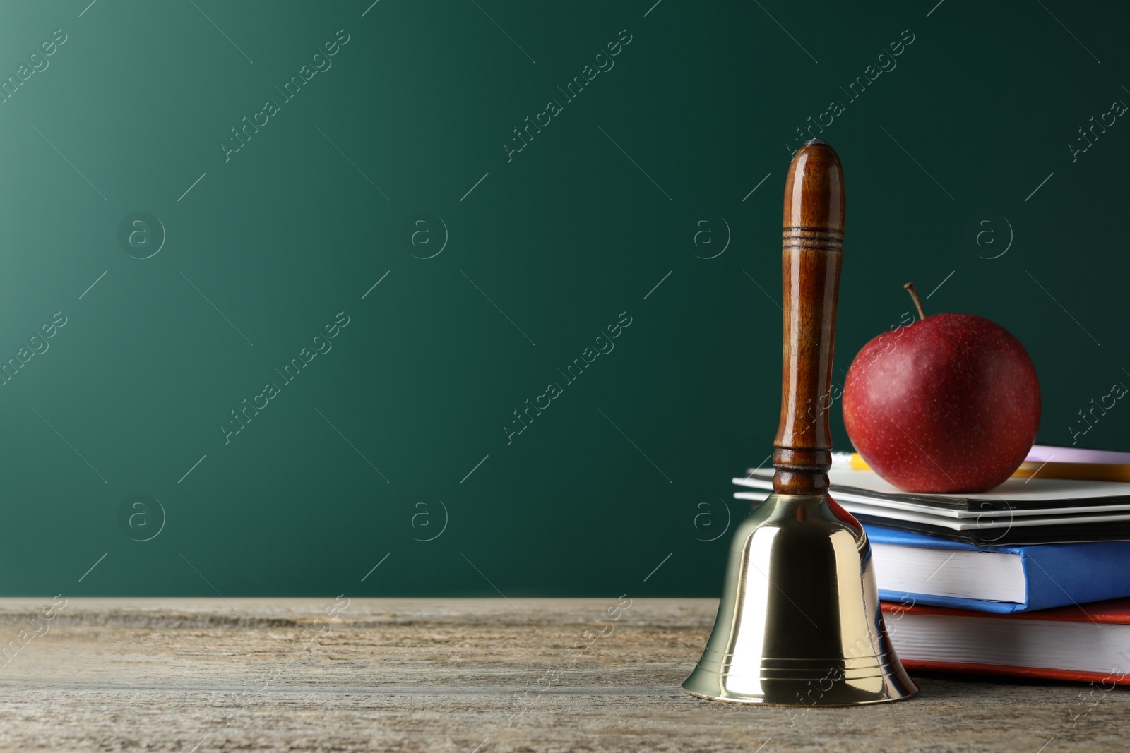 Photo of Golden school bell, apple and books on wooden table near green chalkboard. Space for text