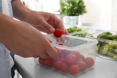 Man putting cherry tomato into plastic container with fresh vegetables in kitchen, closeup. Food storage
