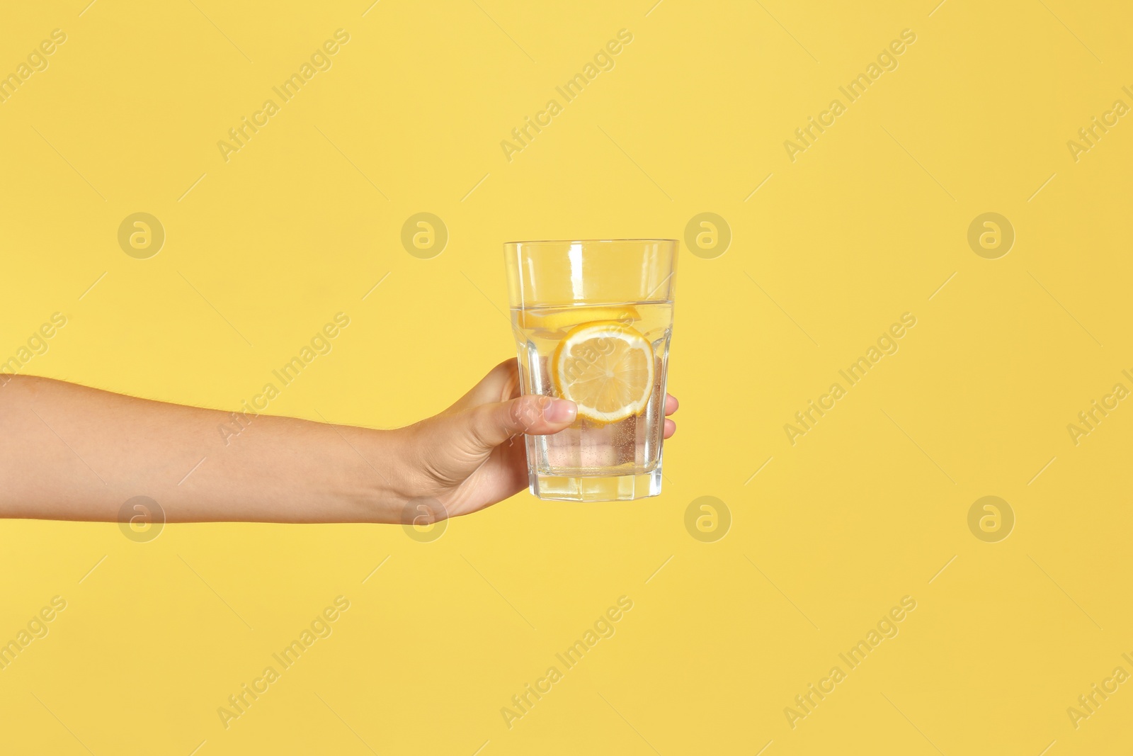 Photo of Young woman holding glass of lemon water on yellow background, closeup
