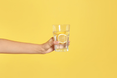 Photo of Young woman holding glass of lemon water on yellow background, closeup