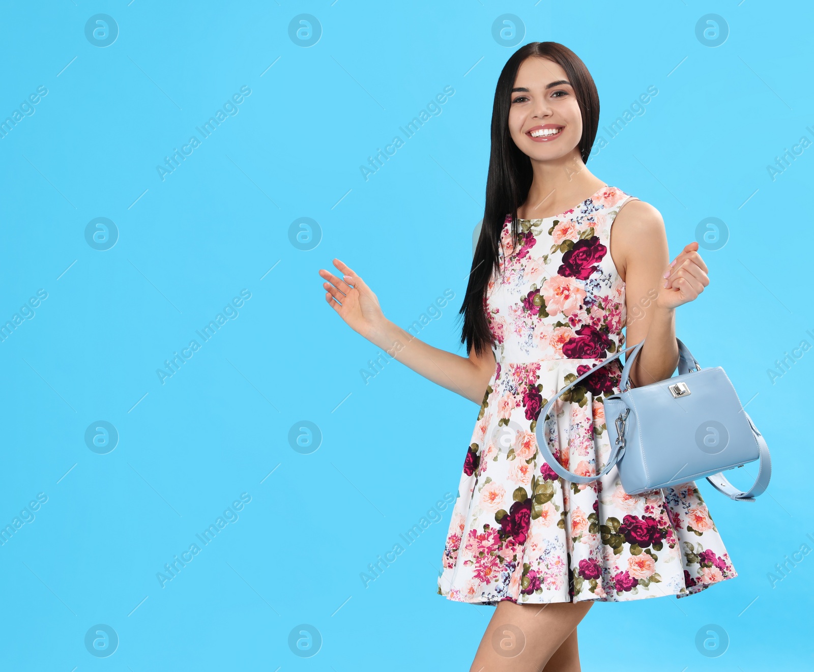 Photo of Young woman wearing floral print dress with stylish handbag on light blue background. Space for text