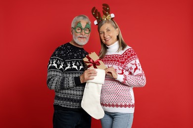 Senior couple in Christmas sweaters taking gift from stocking on red background