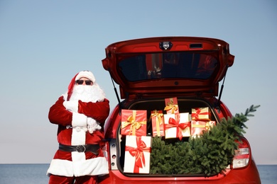 Authentic Santa Claus near car with open trunk full of presents and fir tree at sea