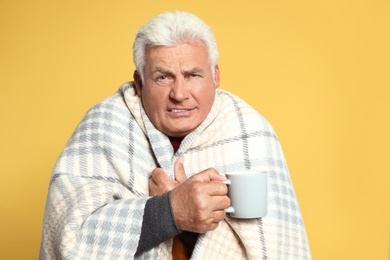 Senior man wrapped in blanket with cup of hot drink on yellow background. Cold symptoms