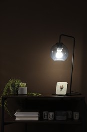 Photo of Stylish lamp, green plants and other stuff on table near brown wall indoors. Interior design