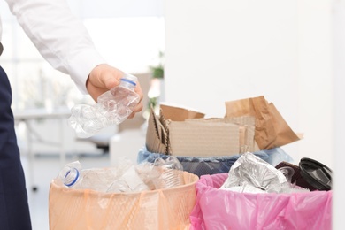 Man putting used plastic bottle into trash bin in office, closeup. Waste recycling