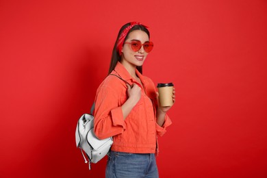 Beautiful young woman with stylish leather backpack and cup of coffee on red background