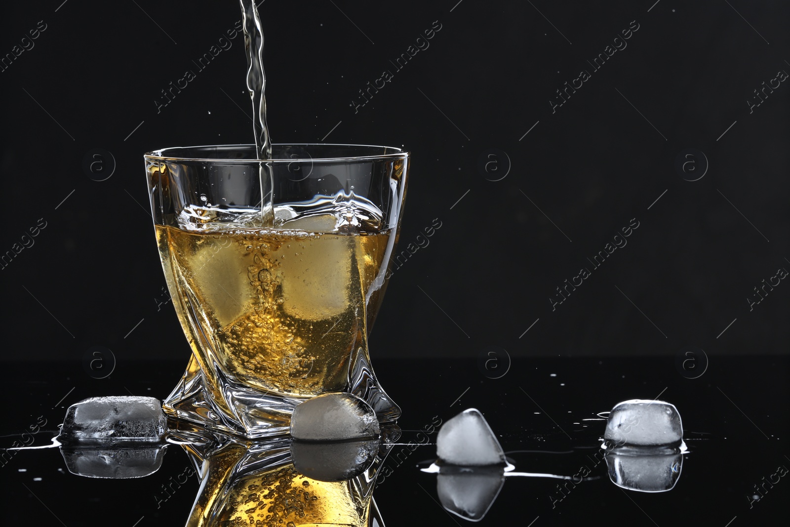 Photo of Pouring tasty whiskey into glass with ice cubes at mirror table against black background, closeup. Space for text