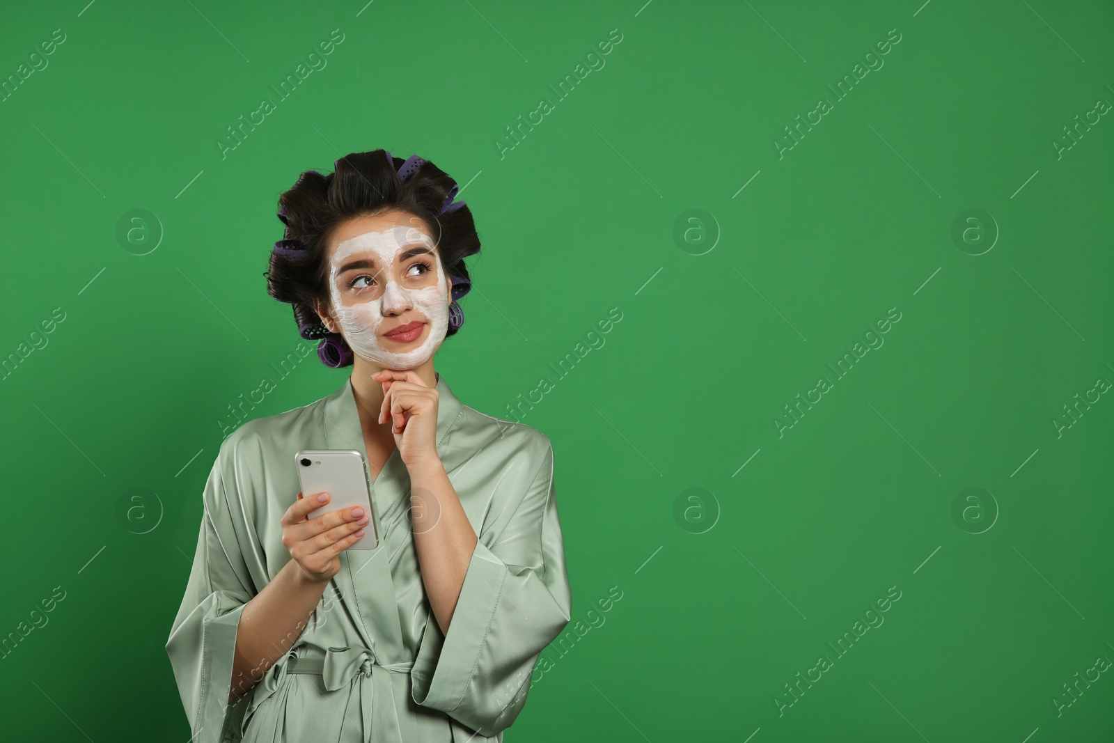 Photo of Happy young woman in silk bathrobe with hair curlers and facial mask using smartphone on green background, space for text