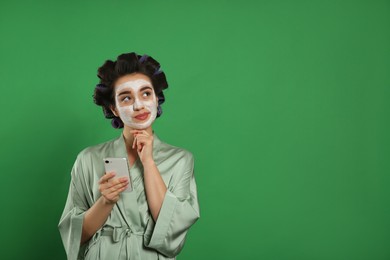 Happy young woman in silk bathrobe with hair curlers and facial mask using smartphone on green background, space for text