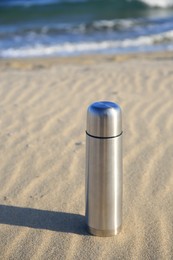 Metallic thermos with hot drink on sand near sea