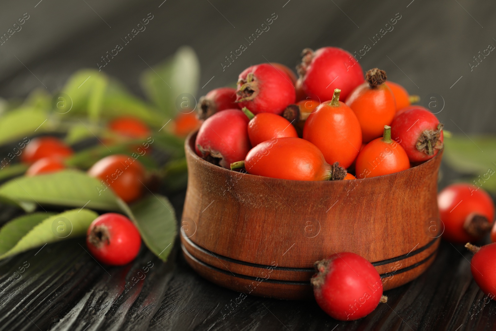 Photo of Ripe rose hip berries with green leaves on black wooden table, closeup