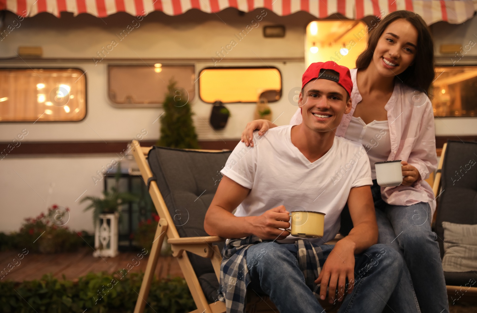 Photo of Happy couple with cups resting on deck chairs near motorhome. Camping season