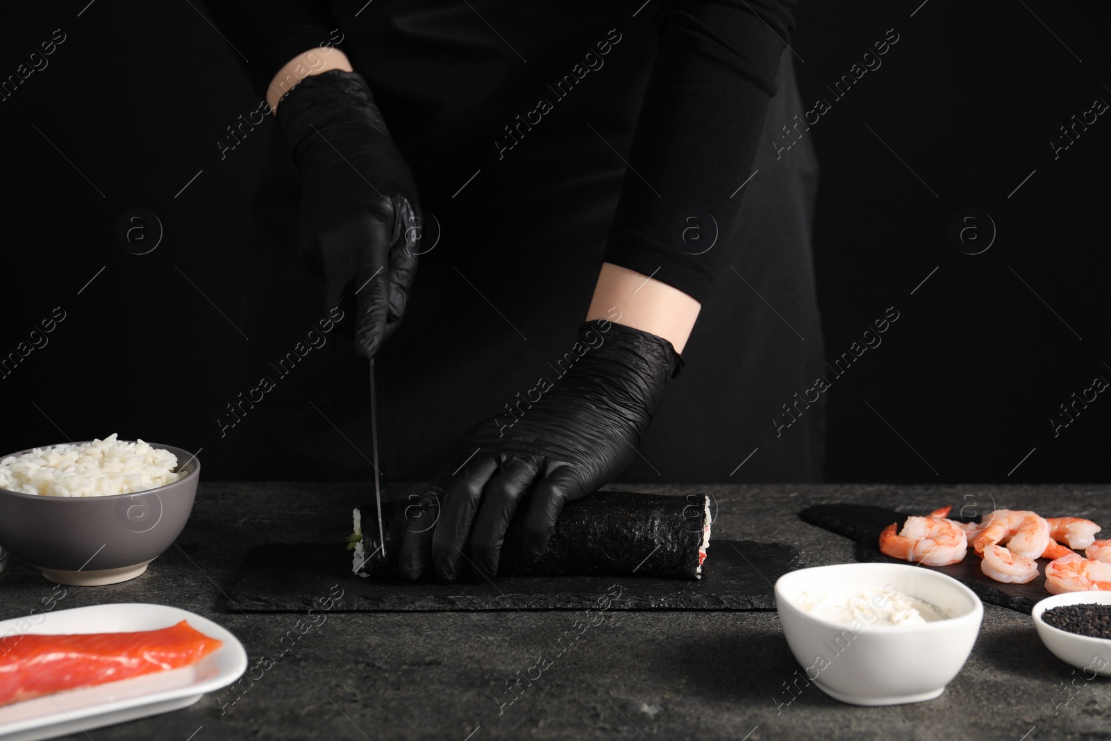 Photo of Chef in gloves cutting sushi roll at dark textured table, closeup