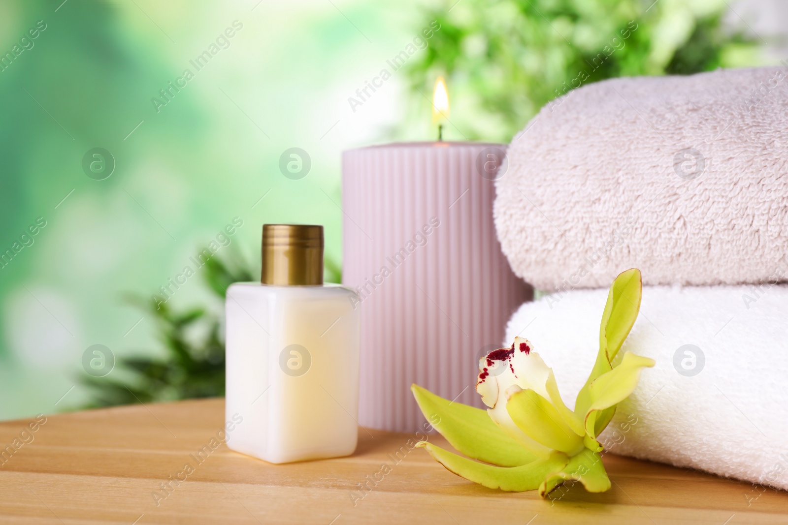 Photo of Lotion, towels and exotic flower on wooden table against blurred green background, space for text