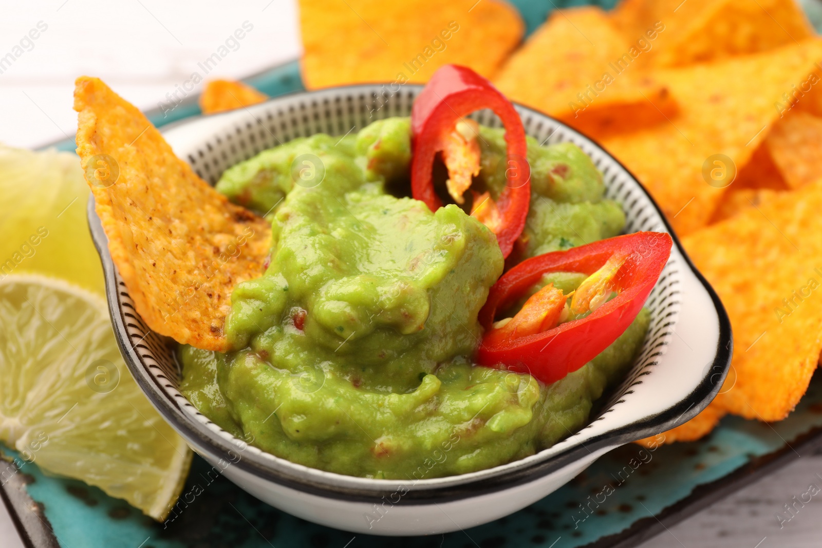 Photo of Bowl of delicious guacamole with chili pepper, nachos chips and lime on plate, closeup