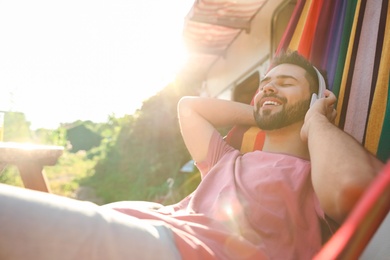 Photo of Young man listening to music in hammock near motorhome outdoors on sunny day