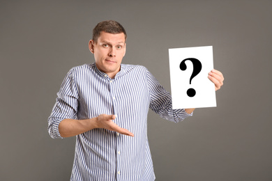 Emotional man holding paper with question mark on grey background