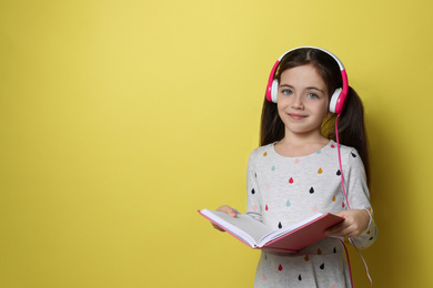 Photo of Cute little girl with headphones listening to audiobook on yellow background. Space for text