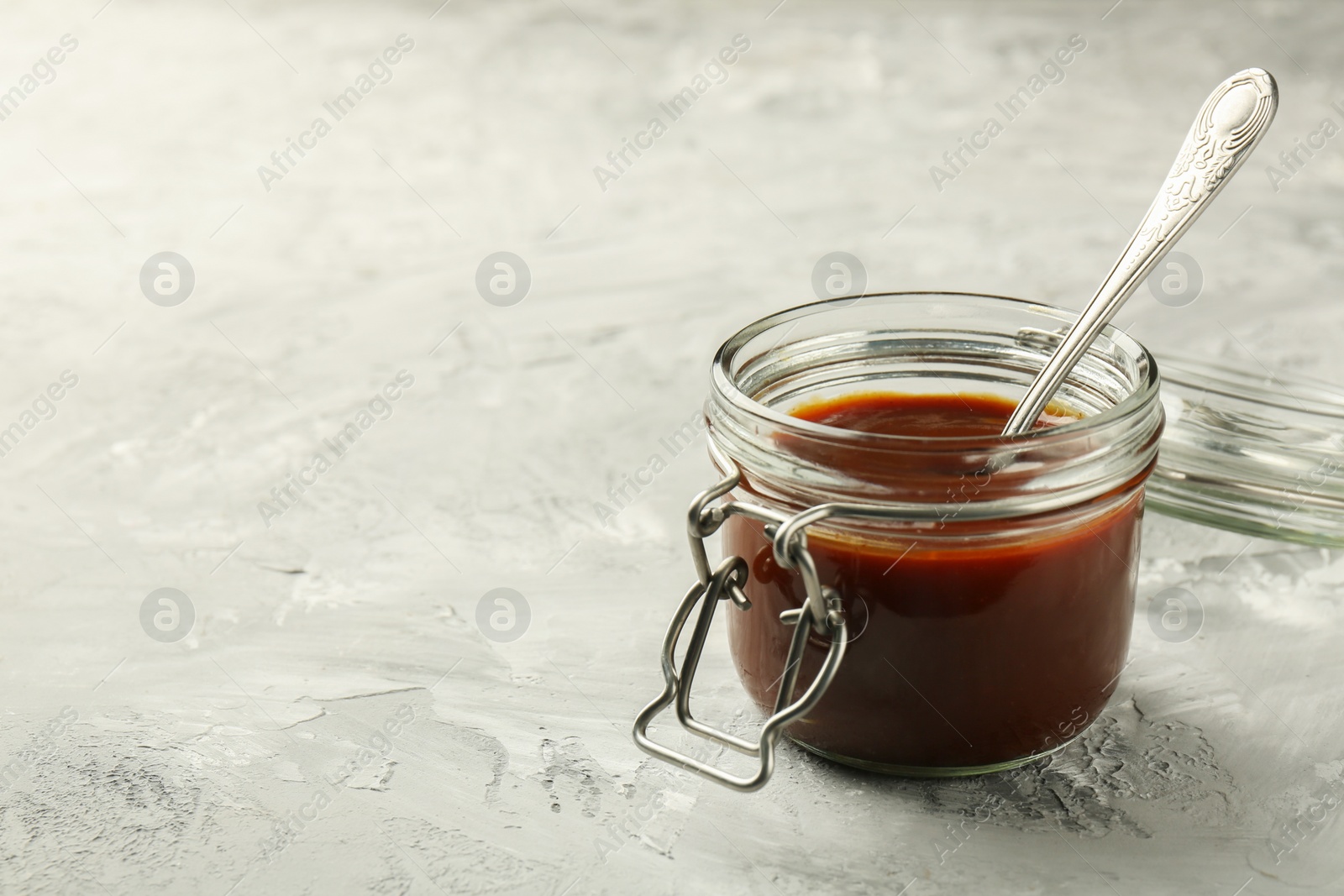 Photo of Tasty barbeque sauce in jar on grey textured table, closeup. Space for text