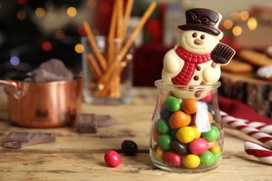 Photo of Funny chocolate snowman candy in glass jar full of dragee on wooden table, closeup. Space for text