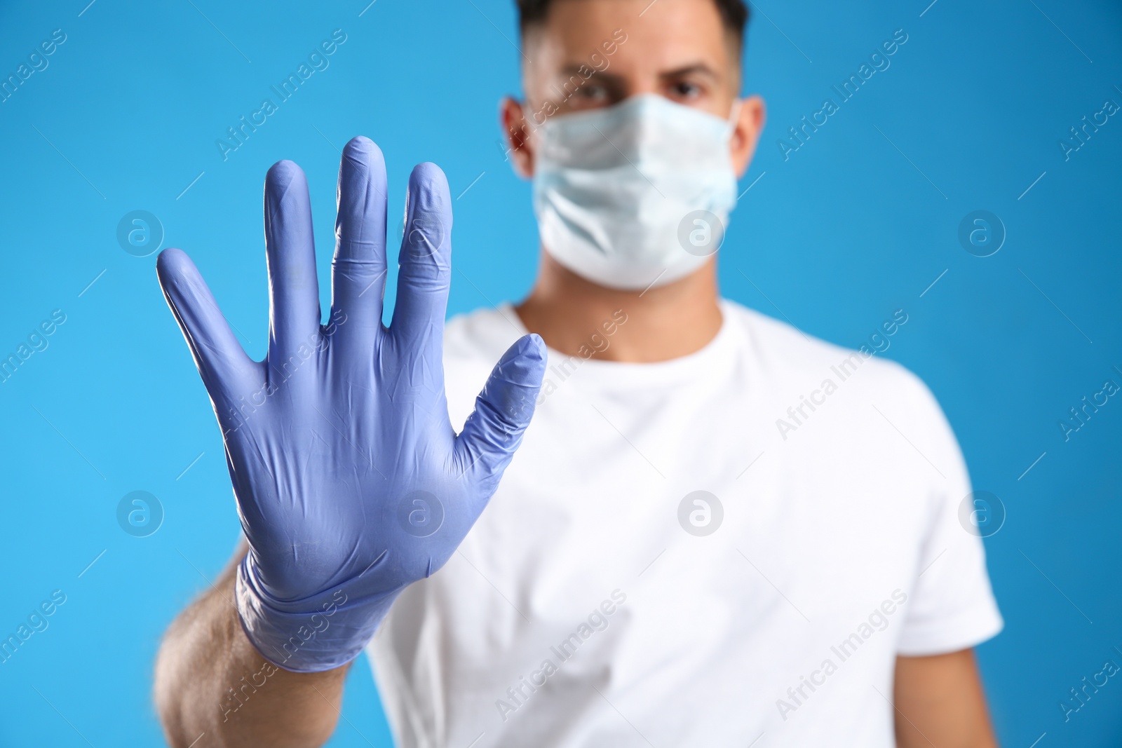 Photo of Man in protective face mask and medical gloves showing stop gesture against blue background, focus on hand