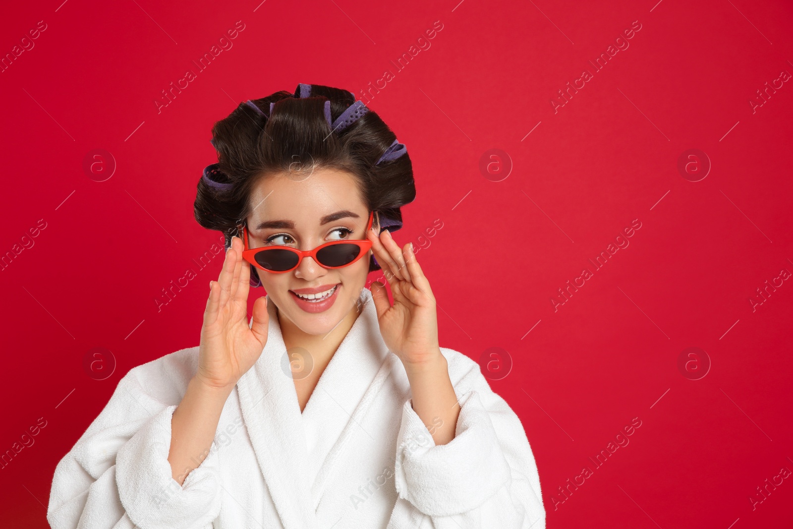 Photo of Happy young woman in bathrobe with hair curlers and sunglasses on red background, space for text