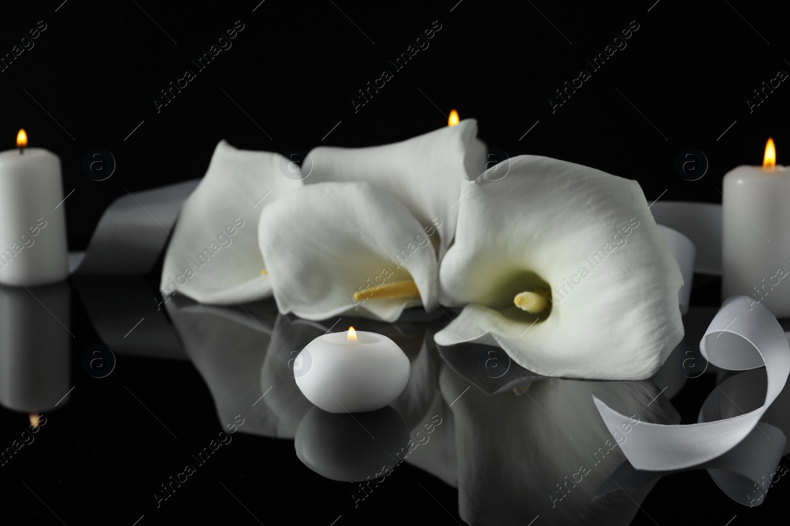 Photo of Burning candles and white calla lily flowers on black mirror surface in darkness, closeup. Funeral symbols