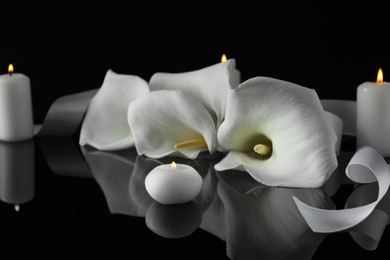 Burning candles and white calla lily flowers on black mirror surface in darkness, closeup. Funeral symbols