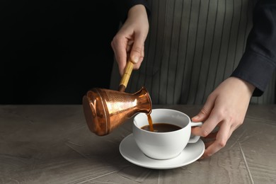 Photo of Turkish coffee. Woman pouring brewed beverage from cezve into cup at gray table against black background, closeup. Space for text