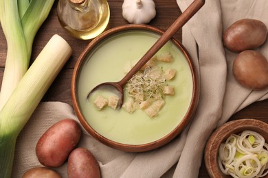 Photo of Bowl of tasty leek soup, spoon and ingredients on wooden table, flat lay